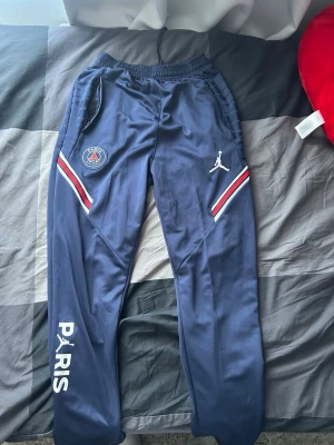 PSG x Jordan blå träningsbyxor - Mörkblå träningsbyxor från PSG x Jordan med vita och röda detaljer längs fickorna, PSG-logga på vänster ben och Jumpman-logga på höger ben. Texten 'PARIS' i vitt längs vänster ben. Resår i midjan och smal passform.