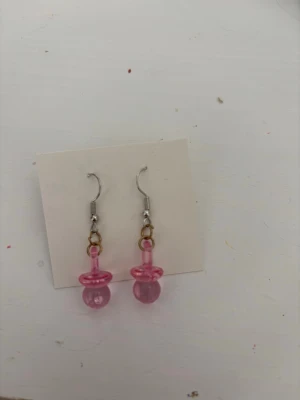 Rosa nappformade örhängen - Säljer ett par unika örhängen i form av rosa nappar. De är gjorda av plast och har krokar i metall. Perfekta för dig som vill sticka ut med en lekfull och färgglad accessoar.