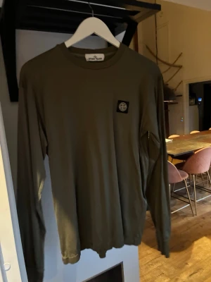 Olivgrön långärmad tröja Stone Island - Snygg olivgrön långärmad tröja från Stone Island med klassisk logotyp-patch på bröstet. Tröjan har rund halsringning och är tillverkad i mjukt bomullsmaterial. Perfekt för en avslappnad och stilren look.