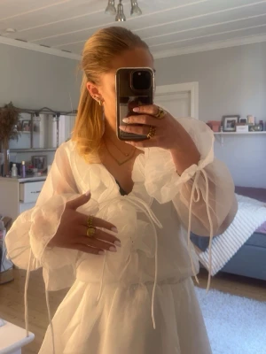 Vit volangklänning med puffärmar - Supersöt vit klänning med volanger och transparenta puffärmar. Klänningen har knytband vid ärmsluten och en kort, flowy kjol med lager på lager-effekt. Perfekt för dig som gillar romantisk och trendig stil. Från Gina tricot, helt oanvänd 