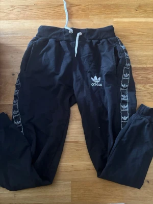 Adidas mjukisbyxor  - Svarta mjukisbyxor från Adidas med vita detaljer och snöre i midjan. Byxorna har ribbade muddar vid bensluten och Adidas logga samt mönstrade band längs sidorna. 