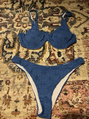 Blå bikini med blommönster - Snygg oanvänd blå bikini med blommigt mönster och vadderad bygel-bh. Bikinin har breda axelband och klassiska bikinitrosor. Toppen är i strl M & trosor i L! ☀️
