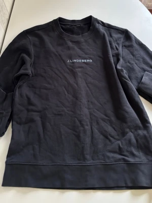 Svart sweatshirt från J.Lindeberg - Snygg svart sweatshirt från J.Lindeberg med diskret logga på bröstet. Tröjan har rund halsringning, ribbade muddar och är tillverkad i mjukt bomullsmaterial. Perfekt för en clean och stilren look.