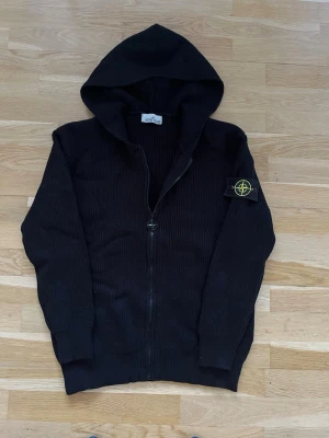 Svart stickad hoodie från Stone Island - Säljer en svart stickad hoodie från Stone Island med dragkedja framtill och klassisk logotyp-patch på ärmen. Tröjan har ribbade muddar och är tillverkad i ett mjukt, grovstickat material. Perfekt för dig som gillar streetwear och vill ha något både snyggt och bekvämt.