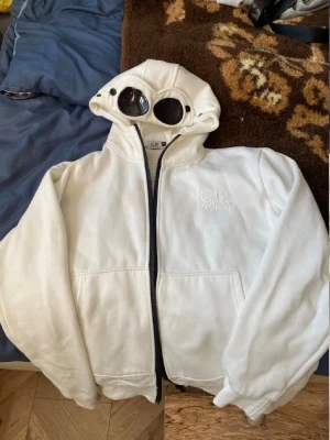 Vit hoodie med goggles från C.P. Company - Säljer en vit hoodie från C.P. Company med ikoniska inbyggda goggles i huvan. Hoodien har dragkedja framtill, två fickor och broderad logga på bröstet. Perfekt för dig som vill sticka ut med en unik streetwear-look.