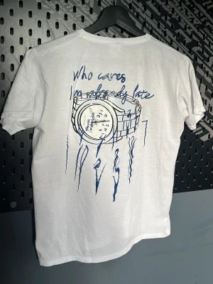 Vit BVALDI t-shirt med blått tryck - Vit t-shirt från BVALDI i bomull med blått tryck på ryggen som föreställer en klocka och texten 'who cares I'm already late'. Framtill finns en liten blå BVALDI-logga på bröstet. Klassisk rund hals och korta ärmar.