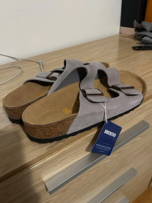 Grå mockasandaler från Birkenstock - Snygga sandaler från Birkenstock i ljusgrå mocka med två justerbara remmar och metallspännen. Fotbädden är anatomiskt utformad och har en bekväm korksula. Perfekta för en avslappnad och trendig stil.