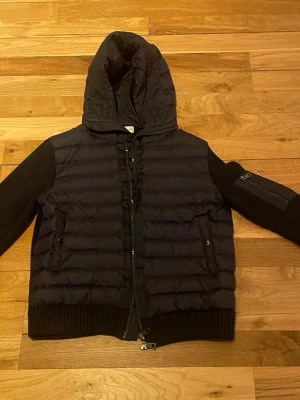 Moncler cardigan - Snygg marinblå cardigan från Moncler med stickade ärmar och huva. Jackan har två fickor fram, ribbade muddar och en Moncler-logga på ena ärmen. Funkar i de flesta väder. Sitter bra och är i väldigt bra skick. Säljer då den inte passar mig längre 