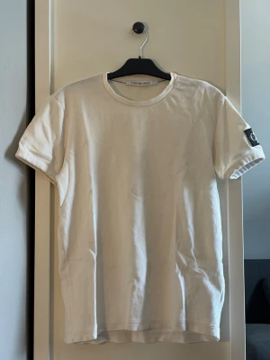 Vit t-shirt från Calvin Klein Jeans - Vit t-shirt från Calvin Klein Jeans med rund halsringning och korta ärmar. T-shirten har en diskret struktur i tyget och en svart logotyp-patch på vänster ärm. Enkel och stilren design som passar till det mesta.