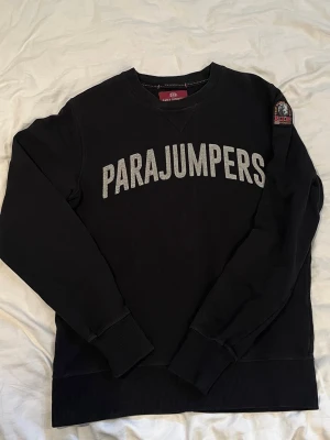 Parajumpers sweatshirt  - Svart sweatshirt från Parajumpers med stor logga i vitt på bröstet och patch på ärmen. Klassisk rund halsringning och ribbade muddar vid ärmslut och nederkant. Perfekt för en avslappnad och stilren look.