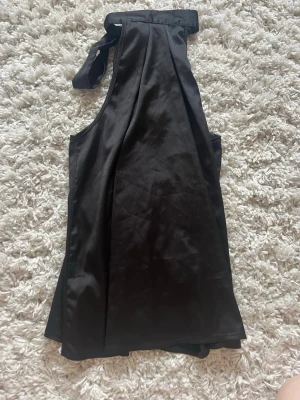 Svart halterneck topp i satin - Snygg svart halterneck topp i glansigt satinmaterial med knytning i nacken. Toppen är ärmlös och har en lös passform som ger en stilren och trendig look. Perfekt att styla med jeans eller kjol för en cool outfit.