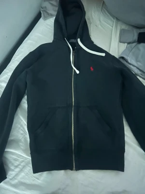 Svart hoodie från Polo Ralph Lauren - Säljer en svart hoodie med dragkedja från Polo Ralph Lauren. Klassisk design med huva och vita snören, samt röd broderad logga på bröstet. Hoodien har kängurufickor och är tillverkad i mjukt material som känns skönt mot huden. Den är storlek XS men sitter bra på S.