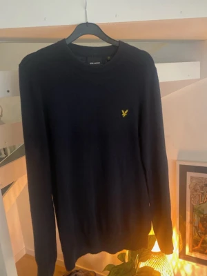 Mörkblå stickad tröja Lyle & Scott - Snygg mörkblå stickad tröja från Lyle & Scott med rund halsringning och den klassiska gula logon broderad på bröstet. Tröjan har långa ärmar och är tillverkad i mjukt material som passar perfekt till lager på lager.
