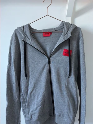 Grå hoodie med dragkedja från HUGO - Snygg grå hoodie från HUGO med dragkedja framtill och svart snörning i huvan. Röd HUGO-logga på bröstet ger en clean men ändå tydlig look. Tillverkad i mjukt bomullsmaterial som är skönt att ha på sig hela dagen.
