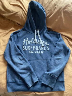 Blå Hollister hoodie med tryck - Mysig blå hoodie från Hollister med vit text 'Hollister Surfboards SO. CALIF' på bröstet. Klassisk känguruficka framtill, vit dragsko i huvan och ribbade muddar. Skriv om du har frågor😊