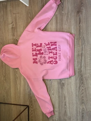 Rosa hoodie med tryck Surf City - Säljer en rosa hoodie med huva och magficka. På ryggen finns ett stort tryck med texten 'MEET ME AT THE OCEAN 1968 SURF CITY' och en hibiskusblomma. Framtill finns en mindre blomma tryckt på bröstet. Perfekt för en avslappnad och cool stil.