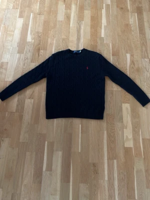 Svart kabelstickad tröja Polo Ralph Lauren - Kabelstickad svart tröja från Polo Ralph Lauren med klassisk röd logga broderad på bröstet. Tröjan har rund halsringning och långa ärmar. Perfekt för dig som gillar stilrena och tidlösa plagg i garderoben.