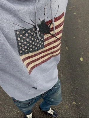 Concreteink hoodie  - Oanvänd Grå hoodie med huva från Concrete ink stor framficka och tryck av amerikanska flaggan med svart färgstänk över bröstet. Mjuk bomullsmix och avslappnad passform, perfekt för en cool streetstil.om ni har några frågor så kan ni kontakta 
