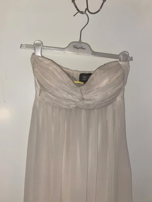 Vit axelbandslös balklänning från Nelly - Elegant ljus långklänning från Nelly med axelbandslös design och draperad överdel. Klänningen har ett vackert fall och en twistad detalj vid bysten. Perfekt för speciella tillfällen, studentfirande och bröllop. Aldrig använd.