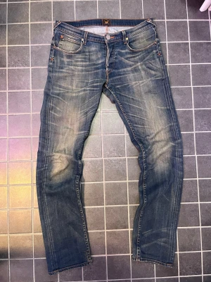 Blå jeans från Lee med slitningar - Snygga blå jeans från Lee med klassisk femficksdesign och coola slitningar framtill. Jeansen har orangea sömmar och en rak passform. Perfekta för en avslappnad och trendig look.