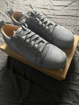 Louiboutin skor - Snygga gråa sneakers från Christian Louboutin i mocka med klassisk röd sula och diskret logga undertill. Skorna har rund tå, gråa skosnören och en stilren design som passar till många outfits.