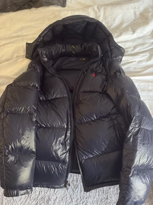 Mörkblå  pufferjacka Polo Ralph Lauren - Mörkblå pufferjacka från Polo Ralph Lauren med huva och dragkedja. Jackan har en glansig finish, breda kanaler och en liten röd logga på bröstet. Perfekt för kalla vinterdagar och riktigt snygg till allt!