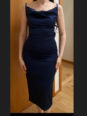 Mörkblå långklänning i satin - Elegant mörkblå långklänning i satin med smala axelband och draperad urringning. Klänningen har en figurnära passform och en stilren, minimalistisk design som ger en lyxig känsla.