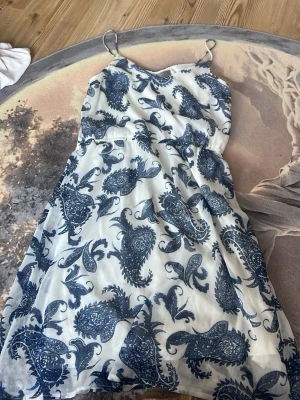 Vit och blå klänning från Vero Moda - Säljer en somrig vit klänning från Vero Moda med blått paisleymönster. Klänningen har tunna axelband, lätt utställd kjol och är tillverkad i ett tunt, luftigt material som passar perfekt för varma dagar.