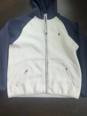 Vit och marinblå hoodie från Polo Ralph Lauren - Snygg hoodie från Polo Ralph Lauren i vitt med marinblå ärmar och huva. Dragkedja framtill, två fickor med dragkedja och klassisk broderad logga på bröstet. Mjuk fleece på insidan för extra komfort. Perfekt för en avslappnad och stilren look.