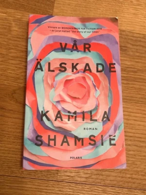 Vår älskade av Kamila Shamsie - Romanen 'Vår älskade' av Kamila Shamsie har ett färgstarkt omslag i rosa, rött, lila och turkost med ett abstrakt blommotiv. Boken är på svenska och utgiven av Polaris. En berättelse om familjeband, kärlek och lojalitet.