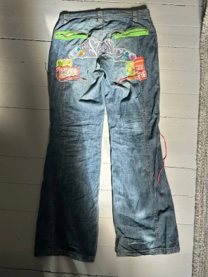 Blå jeans med färgglada lappar - Unika blå jeans med broderade och färgglada lappar, snören och detaljer i neonfärger. Vida ben och låg midja. Perfekta för dig som vill sticka ut med en kreativ och lekfull stil.