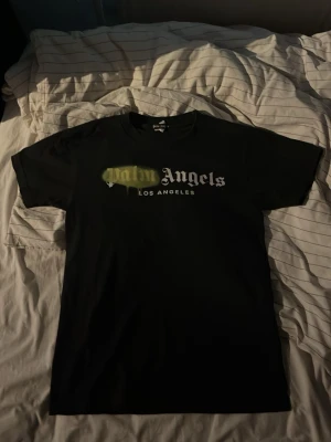Palm Angels svart t-shirt med tryck - Svart t-shirt från Palm Angels med vit logga och texten 'Los Angeles' på framsidan. Unik detalj med grönt sprayliknande tryck över loggan. Baksidan har stor vit Palm Angels-tryck. Klassisk passform och rund hals.