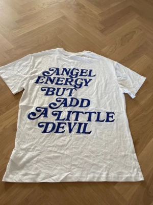 Vit t-shirt med blå texttryck - Vit t-shirt med coolt blått texttryck på ryggen: 'Angel energy but add a little devil'. Mindre tryck på bröstet. Klassisk passform och rund hals. Perfekt för dig som gillar statement-plagg och vill sticka ut lite extra.