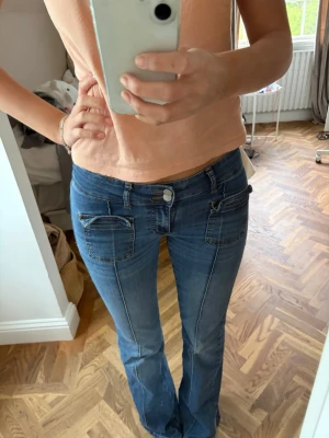 Blå bootcut jeans med fickdetaljer - Säljer ett par blå bootcut jeans med coola fickdetaljer framtill och markerade sömmar. Jeansen har låg midja och dragkedjor på fickorna. Perfekta för dig som gillar en avslappnad men trendig stil.