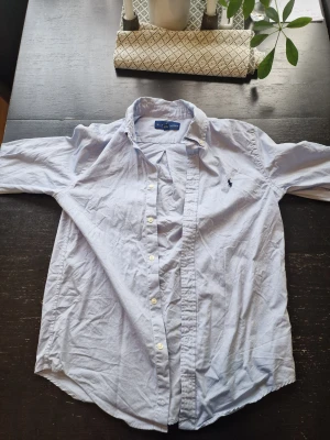 Ljusblå skjorta från Ralph Lauren - Klassisk ljusblå skjorta från Ralph Lauren med button-down krage och broderad logga på bröstet. Skjortan har långa ärmar och vita knappar framtill. Perfekt till jeans eller chinos för en clean och stilren look.