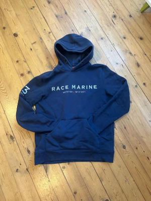 Marinblå hoodie Race Marine - Snygg marinblå hoodie från Race Marine med tryck på bröstet och siffran 83 broderad på ärmen. Klassisk känguruficka framtill och huva. Mjuk bomullsblandning som känns skön mot huden. Perfekt för en avslappnad och sportig stil.