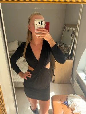 Svart klänning med cut-outs - Snygg svart kort omlottklänning med långa ärmar och djup v-ringning. Klänningen har trendiga cut-outs i midjan och figurnära passform.