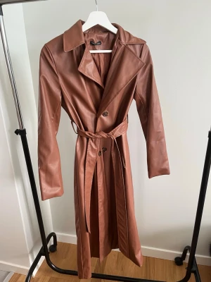 Brun trenchcoat i skinnimitation  - Snygg brun trenchcoat från SHEIN i skinnimitation. Lång modell med klassisk krage, knäppning framtill och matchande bälte i midjan. Perfekt för dig som vill ha en trendig och stilren look med en touch av retro.