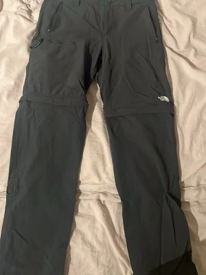 Gråa cargobyxor från The North Face - Gråa cargobyxor från The North Face med dragkedjefickor och avtagbara ben så de kan bli shorts. Byxorna har en sportig look och är tillverkade i ett lätt och slitstarkt material, perfekt för äventyr och outdoor.