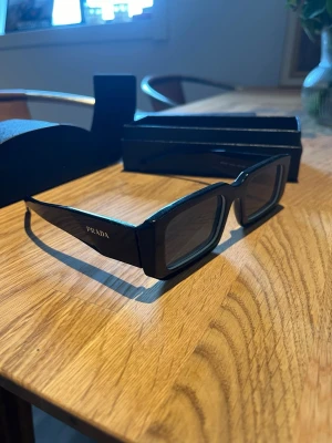 Svarta solglasögon från Prada - Snygga svarta solglasögon från Prada med rektangulära bågar och diskret logga på sidan. Kommer med originalfodral och ask. Perfekta för dig som vill ha en stilren och trendig accessoar till sommaren.