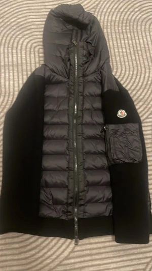 Svart pufferjacka från Moncler - Säljer en svart pufferjacka från Moncler med huva och quiltad front. Jackan har svarta ärmar i mjukt material, två fickor med knapp framtill och en extra ficka med dragkedja på ena ärmen. Klassisk Moncler-logga på ärmen. Perfekt för kalla vinterdagar, våren och hösten också. Scannen fungerar på loggan. 
