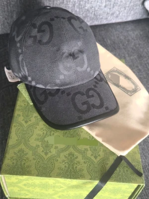 Svart keps med GG-mönster - Säljer en svart kepsi i storlek S (57 cm). Kepsen har det klassiska GG-mönstret i mörkgrått och svart över hela ytan och är tillverkad i ett slitstarkt tygmaterial. Skärmen har en snygg svart kant och kepsen kommer med dustbag och box. Oanvänd! 