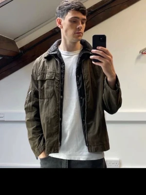 Olivgrön overshirt med dragkedja CP company - Snygg olivgrön overshirt med dragkedja framtill och flera fickor, bland annat på bröstet och ärmen. Jackan har en avslappnad passform och är tillverkad i bomull, perfekt för lager-på-lager-stil.
