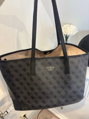 Svart monogramväska från Guess - Snygg svart handväska från Guess med klassiskt monogrammönster och guldfärgade detaljer. Väskan har två långa axelband och rymligt innerfack, samt en mindre matchande väska med dragkedja. Tillverkad i konstläder med stilren design.🤍