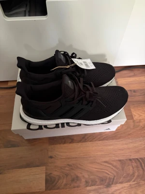 Adidas UltraBoost svarta sneakers - Svarta Adidas UltraBoost sneakers med stickad ovandel och klassiska tre ränder på sidan. Skorna har en vit Boost-sula för extra komfort och svart yttersula. Perfekta för dig som gillar sportig och stilren look.