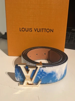 Louis Vuitton blått bälte med LV-spänne - Snyggt bälte från Louis Vuitton i blått och vitt med molnliknande mönster. Bältet har ett stort, guldigt LV-spänne och är gjort i läder. Perfekt accessoar för att lyfta din outfit.