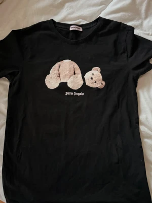 Svart Palm Angels t-shirt med björn - Svart t-shirt från Palm Angels med ikoniskt björnmotiv på bröstet och stor logga på ryggen. Klassisk passform och rund halsringning. Perfekt statement-plagg för dig som gillar streetwear. Liten i storlek!!!