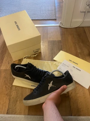 Svarta Axel Arigato sneakers med fågel - Snygga svarta sneakers från Axel Arigato med vit fågelbrodyr på sidan och vit sula. Skorna har klassisk låg profil, svarta skosnören och är tillverkade i slätt läder. Perfekta för dig som gillar stilrena och trendiga sneakers. Storlek 41 passar 42 Box medföljer ej 