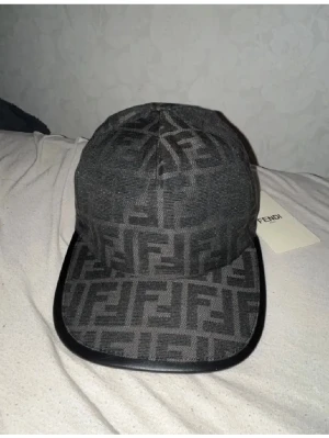 Grå keps från Fendi med FF-mönster - Snygg grå keps från Fendi med det klassiska FF-logomönstret över hela kepsen. Kepsen har böjd skärm med svart kant i läderimitation och justerbar rem baktill. Perfekt accessoar för att lyfta din streetwear-look.
