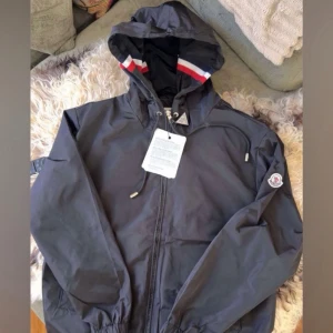 Svart vindjacka från Moncler - Snygg svart vindjacka från Moncler med huva och dragkedja framtill. Jackan har justerbar dragsko i huvan och Moncler-logga på ärmen. Fodret i huvan har röd, vit och blå rand. Perfekt för blåsiga dagar.